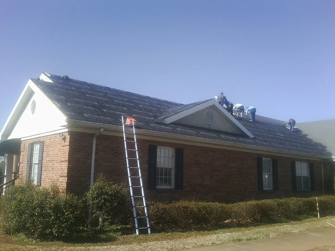 Crew tearing shingles off a brick ranch roof with a ladder set — Emergency Roof Repair work by our crew in Farner