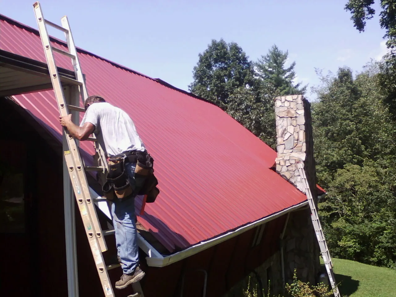 Roofer on a ladder beside a red metal roof and stone chimney — Soffit & Fascia Repair by Roofing Brothers in Farner